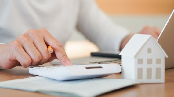 Eine Person benutzt einen Taschenrechner und überprüft Dokumente auf einem Schreibtisch, neben einem kleinen Modellhaus.