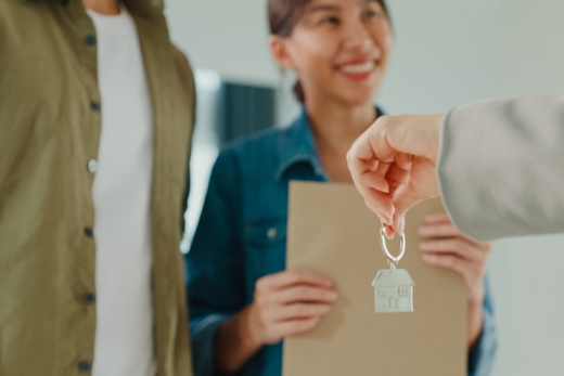 Eine Person übergibt einer anderen einen Schlüssel, während im Vordergrund ein Klemmbrett mit Dokumenten liegt.